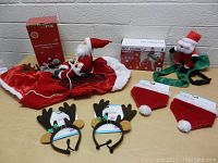 Overview of all dog Christmas costume items including 2 Ride Along Santa costumes visible with packaging, 2 reindeer headbands and 2 bandannas laid out on surface.