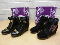 Two pairs of women's wedge boots displayed side by side with boxes behind them. One pair is black leather with gold accents and the other pair is black with floral panels and decorative elements.