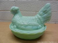 Vintage pale green glass hen sitting on a nest covered dish shown assembled on a flat surface with a brick wall background.