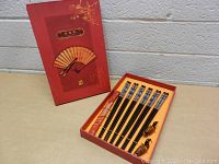 Photo showing boxed set containing 6 chopsticks and 2 wooden bird holders in a red decorative box with fan motif on lid.