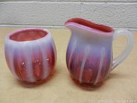 Pair of hand blown glass vase and pitcher showing red to purple gradient with vertical white patterns, on a beige table against cinder block wall