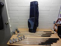 Full view of complete set of golf clubs laid on a table next to a navy blue golf bag standing upright.