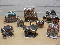 View of 8 assorted ceramic Christmas village houses with snowy roofs on wooden surface