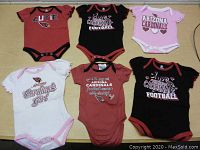 Top view photo of six Arizona Cardinals baby girl bodysuits laid out on a flat surface, showing front designs and colors.