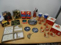 Photo showing assorted Christmas items on table including glass cups, mugs, figurines, candle holders, string lights, napkins, note cards, and plates