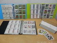 Photo of three binders open to pages showing multiple NFL football cards and a box of loose cards and cards spread out on the table.
