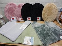 Photo showing two pink, two black, and one beige oval toilet seat covers, plus light gray and light green rectangular bath rugs, all new and plush.