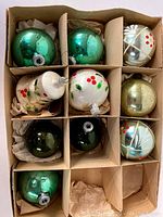 View of ten vintage Christmas balls in original cardboard box with paper padding. Mixed green, white, and gold colors. Some painted with holly berry motifs.