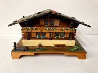 Front and side angle of the vintage Swiss chalet style wooden music box showing detailed floral decorations, windows with shutters, bench, and painted flowerbeds.