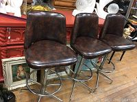 Three vintage swivel bar stools side by side with dark brown vinyl seats and backs on chrome metal bases. They are shown on a wooden floor in a retail setting surrounded by other furniture and decor items.