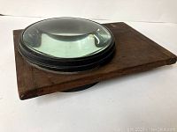 Side angle showing the thick convex glass lens in a black metal frame mounted on a rectangular wooden base with visible wood grain and minor surface wear.