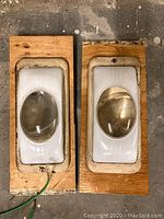 Pair of vintage wall lights mounted on wooden boards on a concrete floor. Both lights have white metal bodies with oval translucent lenses, show wear and aging.