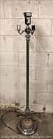 Full view of vintage floor lamp with tall metal pole, three candle-style bulb holders around a central oil reservoir, standing against a concrete block wall.