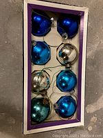 Box containing eight vintage Christmas glass balls in blue, silver, teal colors on cardboard base with purple border.
