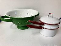 Green enamelware colander with handles and white interior alongside white and red double boiler pot with lid and red handle