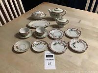 Photo showing full set of tea ware items arranged on a table including plates, oval dish, teacups, creamer, sugar bowl, and teapot, all featuring floral and vine decorative patterns and gold accents.