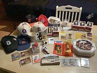 Photo showing assortment of sports memorabilia including hats, baseball and basketball figurines, collector plates, and cards arranged on floor and table.