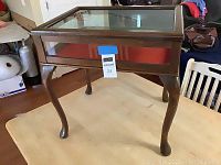Front angled view of vintage wooden curio display table with glass top and red interior lining on wooden legs.