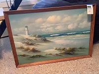 Full view of the framed oil painting depicting beach sand dunes, grasses, lighthouse, and ocean under cloudy sky.