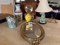 Overview of assorted home decor items on table including marble and pewter wine bottle holder, GMT world wall clock, green porcelain bunny lamp, colorful Porcelanas Guillen vase, yellow smiley sugar bowl, and vintage gold-rimmed oval mirror