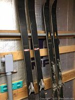 Two pairs of skis standing upright, showing tops and bindings, against a wall with visible piping and structure.