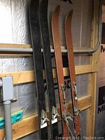 Two pairs of vintage long skis leaning against a wall in a storage area. One pair is black Salomon skis, the other pair is reddish-brown Silveretta skis.