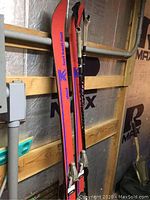 Two vintage Hart-Kash short skis leaning against a wall, showing the bright reddish-orange tops with branding and attached ski poles.