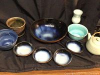 Front view showing variety of ceramic pottery items including cobalt blue bowls, pale turquoise bowl, teapot, goblet, and flower pots arranged on a cloth.