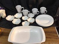 Top view photo of blue Le Creuset ceramic casserole dish, white ceramic Sonoma and Royal Copenhagen plates, multiple floral small bowls, and four white mugs on dark cloth background.