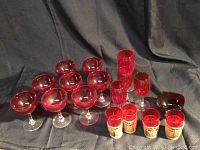 Photo showing 8 red champagne coupe glasses, 6 parfait glasses with colored stems, multiple small red cordial glasses, all arranged on black fabric backdrop.