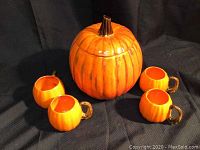 Photo showing one large pumpkin cookie jar and four matching small pumpkin mugs on a dark cloth background.
