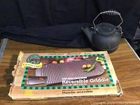 Reversible cast iron griddle in original box with cast iron kettle on top.