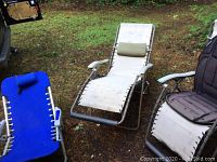 Four outdoor folding chairs on grass with vehicle in background. Two tan recliners, one with black Homemedics back pad, and one blue chair.