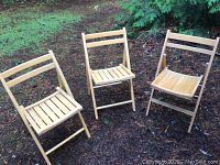 Three blonde oak folding chairs with slatted seats and backs arranged outdoors on soil, showcasing front and slightly top views.