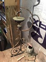 Front angle of ornate metal oil lamp stand and smaller kettle stand with kettle on floor against wall.