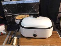 GE 18 Quart Roaster Oven with lid, displayed with its original box, on a wooden surface with four knives beside it.