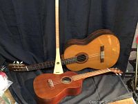 Three stringed instruments grouped together on black backdrop: Fender guitar, Hohner ukulele, and handcrafted long neck instrument.