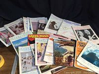 Collection of about 20 vintage Sheet Music magazines stacked and laid out on a surface against a black background.
