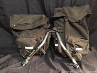 Pair of used black Novara bike panniers with front zip pockets, top flap closures, and Scott speed bar handlebar brace lying on top