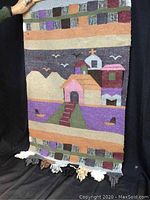 Photo of woven wall hanging showing village scene with houses, mountains, birds, and patterned borders with tassels at bottom