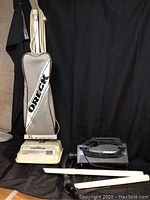 Image shows two Oreck vacuum cleaners: a beige upright with fabric bag and XL Access L-e label, and a black handheld vacuum. Two white extension tubes and a black hose attachment are placed in front.