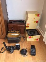 Lot items arranged on floor showing camera bags, boxes of Kodak slide trays, Minolta camera, flash and small camera.