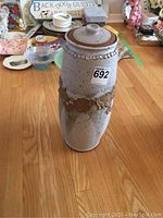 Full view of pottery piece standing on floor with lid on, showing height and applied decorative band.