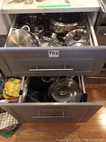Open kitchen drawers showing assortment of stainless steel pots, pans, and glass lids inside two drawers, some pots labeled with brands Lagostina and Meinekuche.