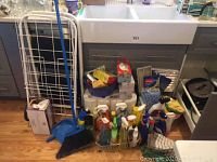 Photo showing front view of entire lot: white metal folding cart, blue broom/dustpan, cleaning sprays, bleach, paper towels, tissues and sponges all grouped together on wooden floor in kitchen.