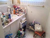 Photo showing bathroom vanity shelf and cabinet with multiple Avon skincare bottles, cleaning supplies under sink, toilet paper and small stools on floor with wall clock on stool.