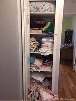 Closet shelf with stacked folded linens including white sheets, patterned fabrics, and blankets.