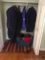 Closet view showing two hanging men's winter coats, snow pants, and plastic tote with scarves and mitts on floor.