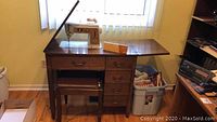 Singer sewing machine placed on wooden sewing desk with drawers and attached fold-out top surfaces, matching cushioned stool below desk.