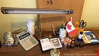Desk area showing task lamp, calculator, tape, calendar, scissors, batteries and other small office tools on wooden surface with corkboard behind.